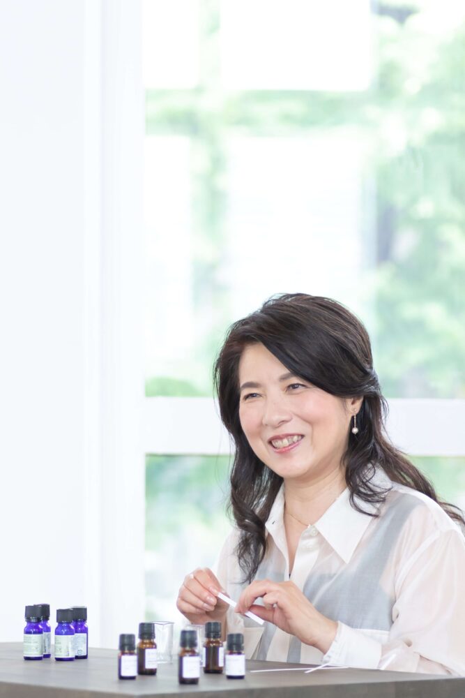 窓辺のテーブルで精油の小瓶を前に微笑む田原範子